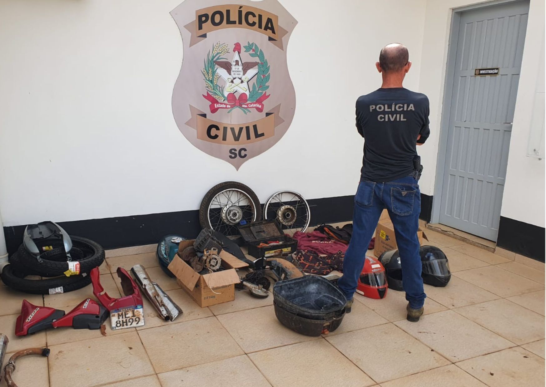 um policial civil em frente a uma parede com a insígnia da policia civil e no chão rodas, capacetes e outras partes de um veículo