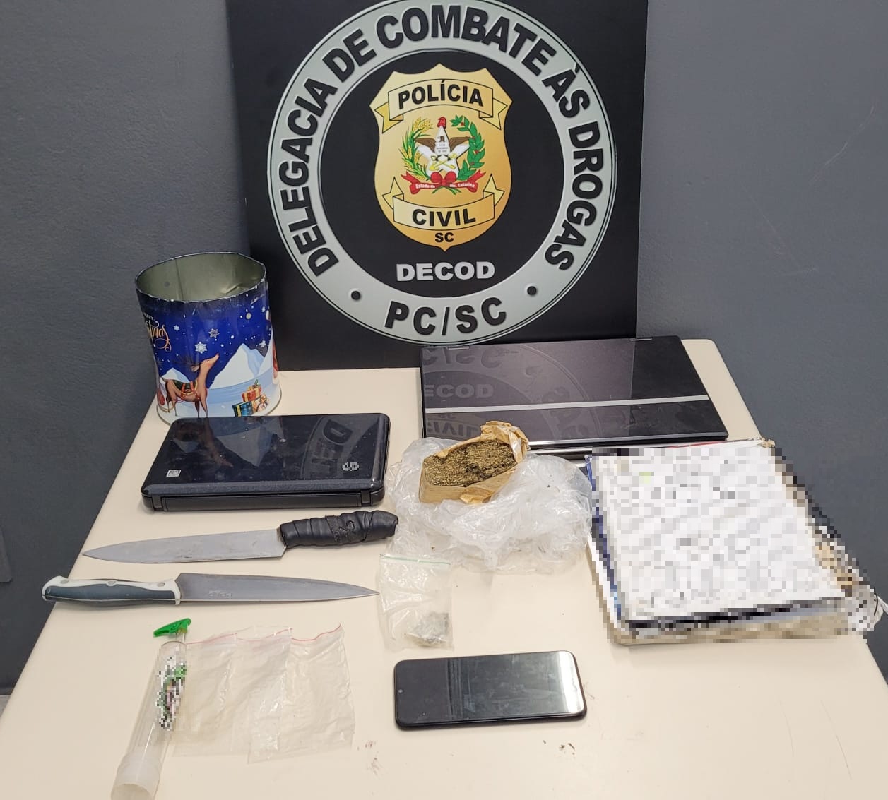 banner da delegacia de combate as drogas com a insígnia da PC, abaixo uma mesa com caderno, duas facas, dois notebooks, droga e plásticos