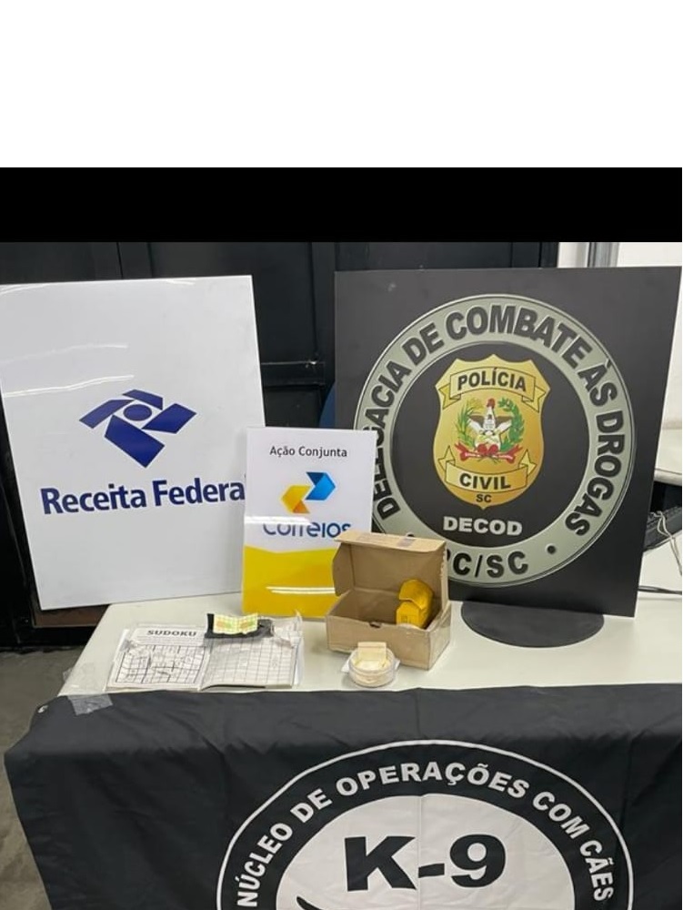 mesa com banner da Receita Federal, Correios e Delegacia de Combate às drogas junto a estes um pacote e faixa Coordenadoria de Operações Policiais com Cães