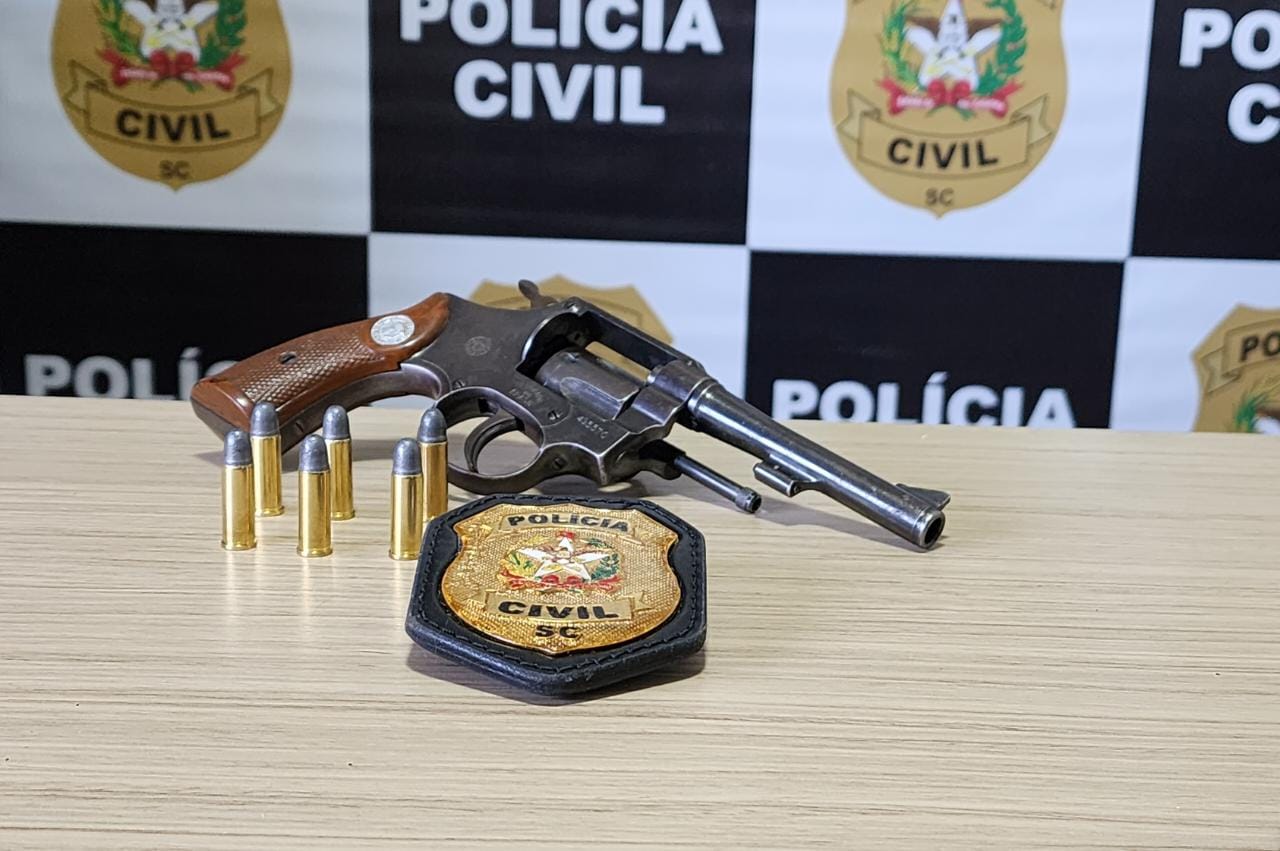 banner da policia civil atrás e na frente sobre uma mesa uma arma, balas e o distintivo da Policia Civil