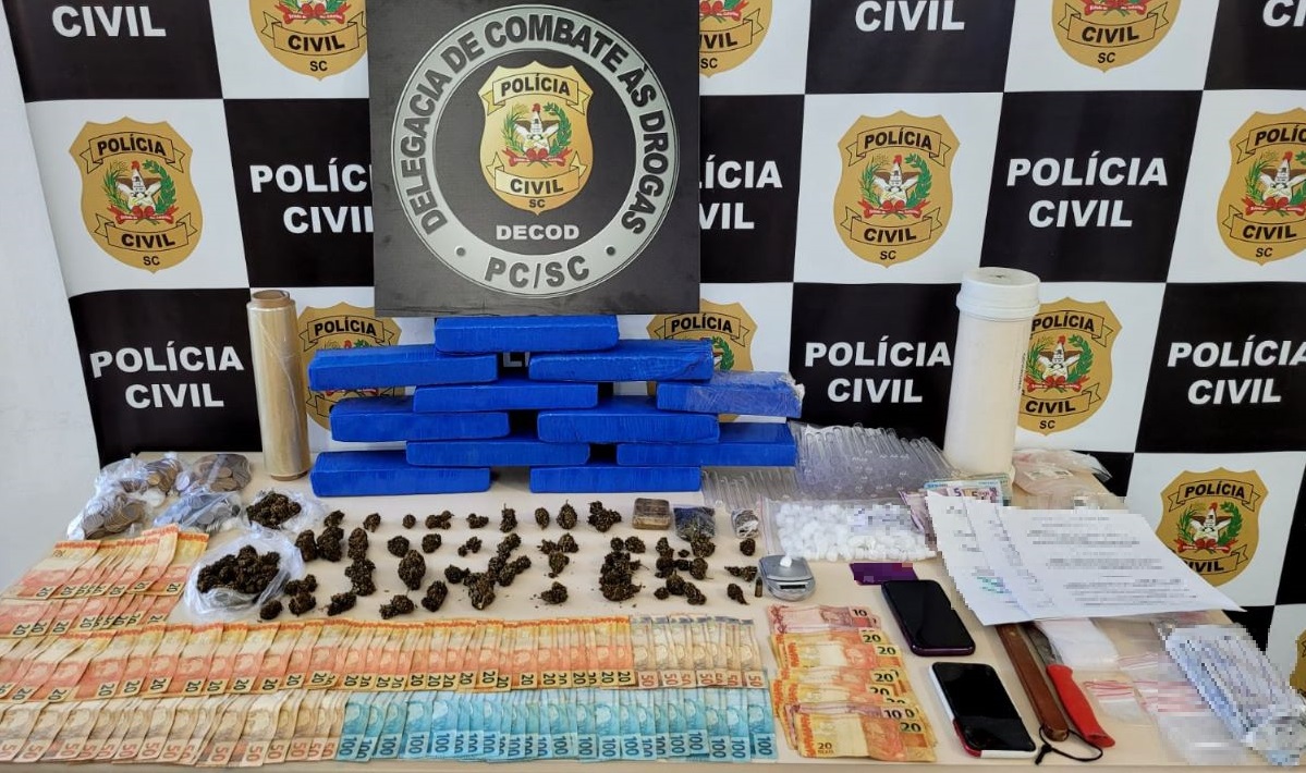banner ao fundo na mesa poster da delegacia da Polícia Civil de combate as drogas com drogas, dinheiro, cartões, celulares, facas e embalagens recolhidas