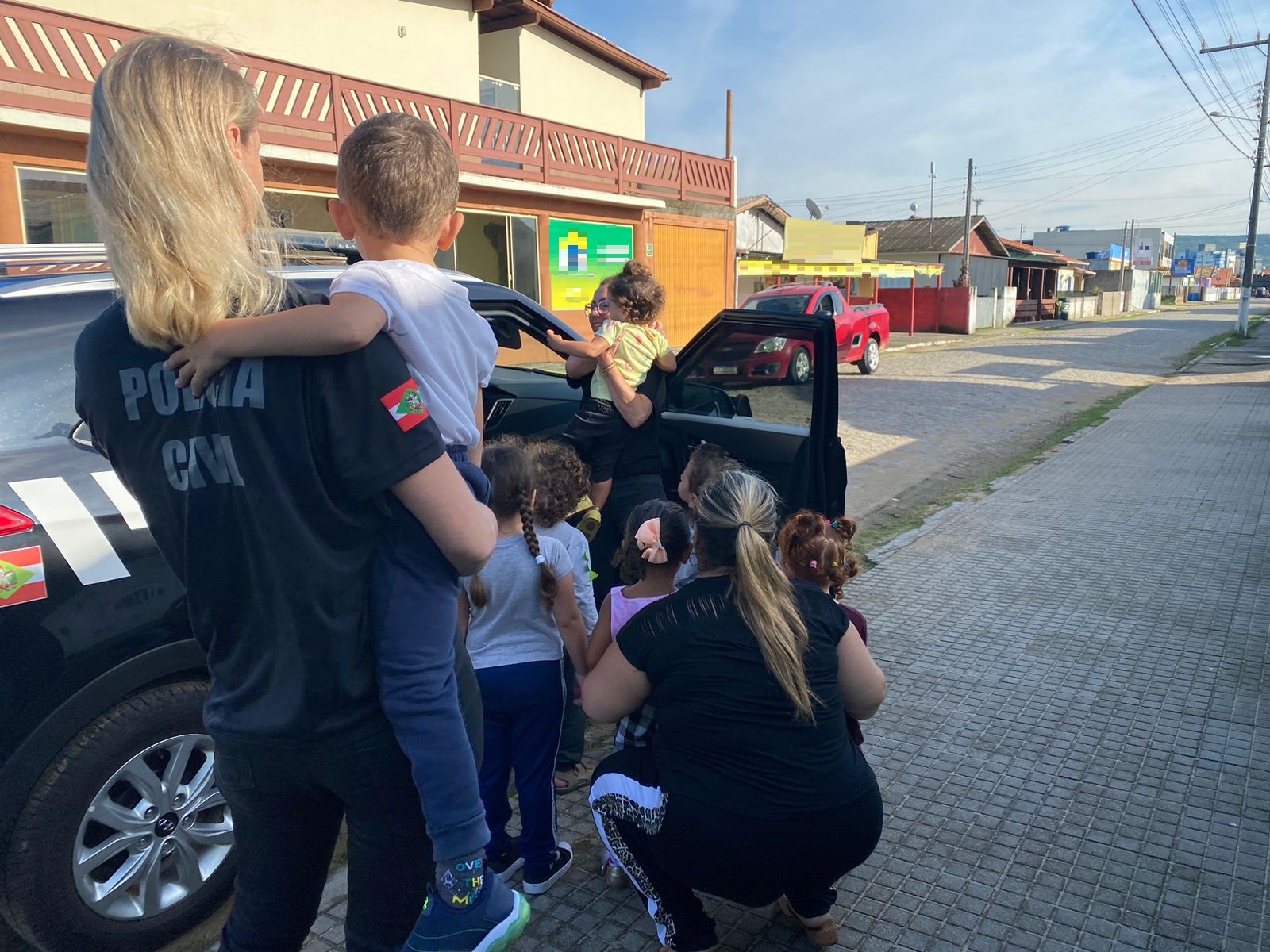 duas policiais civis segurando criança no colo em frente a uma viatura com a porta aberta aonde outras seis crianças acompanhadas por uma terceira policial entram na viatura